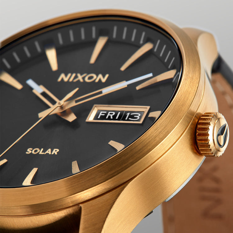 A close-up of a gold Nixon Sentry Solar watch with a black face, gold hour markers, and a date display showing "FRI 13".