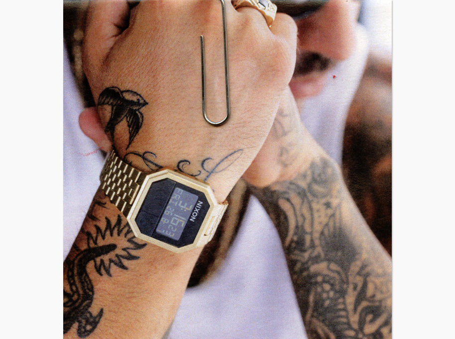 A close-up lifestyle shot of a person's tattooed wrist wearing a gold-tone digital watch with a gold link bracelet. The watch has a black face with a blue digital display showing the time 5:00.