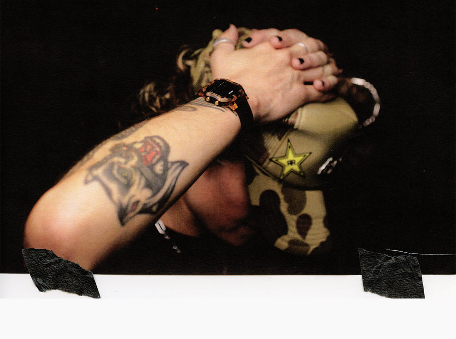 A close-up lifestyle shot of a person with tattoos covering their arms, wearing a camouflage baseball cap with a yellow star logo and a black watch, set against a dark background.