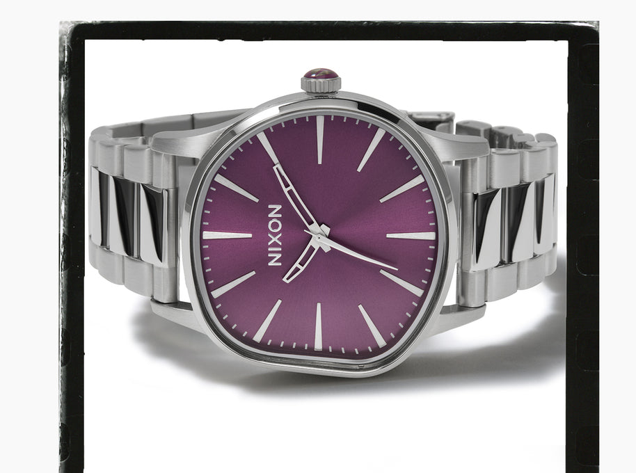 A Nixon wristwatch with a silver-tone metal bracelet and case, featuring a purple dial with silver-tone hour markers and hands.