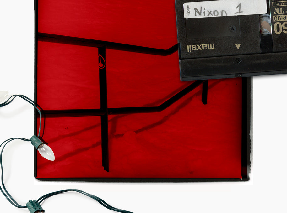 A square red canvas or panel with black geometric line art, photographed on a white background. A black Maxell tape sits on the upper right corner of the artwork, and a string of Christmas lights is draped along the lower left edge.